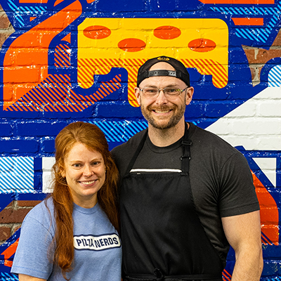 Brent and Katelyn at their pizza shop, in front of a painted mural